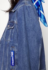 Džínová bunda v světle modrém odstínu, s klasickým límcem, dvěma předními kapsami a modrou značkovou nášivkou na boku. Modrý šátek s potiskem jako doplněk.