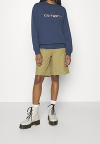 Sudadera marina con el logo "carhartt", pantalones cortos caqui con bolsillos laterales, y botas blancas con cordones y suela negra gruesa.