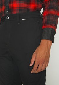 Personne portant un pantalon noir G-Star et une chemise à carreaux rouge et noir avec des poignets foncés, main reposant à côté de la cuisse.
