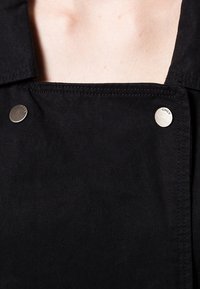 Tessuto denim nero con colletto strutturato, caratterizzato da bottoni a pressione in acciaio. Dettagli di cucitura visibili e una texture liscia.