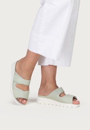 Feet wearing light green open-toe slip-on sandals with white soles, paired with cropped white pants against a plain background.