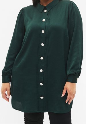 Vestito camicia verde scuro a maniche lunghe con bottoni decorativi bianchi a forma di fiore sul davanti e sui polsini, indossato sopra pantaloni neri.
