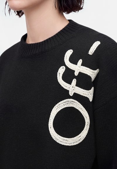 Gros plan d'une personne portant un pull en tricot noir avec de grandes lettres blanches texturées disposées verticalement sur la zone des épaules.