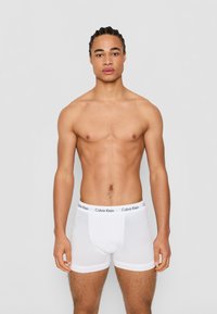 Biele bavlnené boxerky Calvin Klein s čiernym logom na pásku, priliehavý strih a hladký povrch, model predvádzaný s nahým trupom.