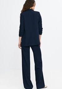 Kobieta z długimi kręconymi włosami, ubrana w granatowy garnitur z podwiniętymi rękawami, stojąca tyłem na białym tle.