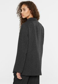 Dark gray blazer in textured fabric with a tailored fit, featuring a notched collar, long sleeves, and a straight-cut silhouette.