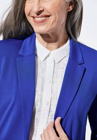 Blazer bleu en tissu lisse, associé à une chemise blanche texturée à boutons, avec des fines rayures et un col classique.