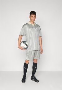 Equipamento de futebol cinzento com padrão geométrico, camisola de mangas curtas, colarinho bege e calções a condizer. O modelo segura uma bola de futebol preto e branco.