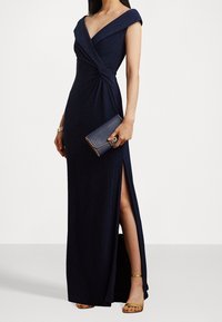Nachtblaues Maxikleid mit schulterfreiem Design, verdrehtem Vorderteil und schulterhohem Schlitz, kombiniert mit einer Clutch und goldenen Riemchensandalen.