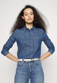 Camisa de ganga azul-escura com um design de botões, bolso no peito e mangas enroladas, combinada com jeans de cintura alta azul-clara e um cinto branco com tachas.