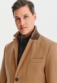Jasnobrązowy wełniany płaszcz z podniesionym kołnierzem, zapięciem na guziki i kieszenią na piersi, noszony na czarnej pikowanej kurtce.