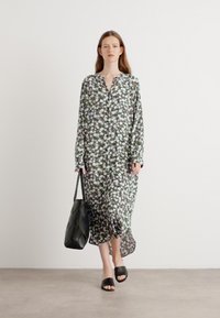 Abito lungo floreale in nero, verde e bianco, con colletto e vestibilità ampia. Abbinato a sandali neri a scivolo e una borsa texturizzata.