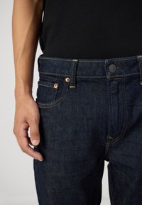 Tumši zili džinsu bikses ar pieguļošu dizainu, piecām kabatām, vara kniedēm un kontrastējošām dzeltenām šuvēm. Tuvplāns uz jostasvietas un roku.