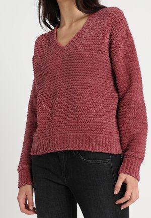 Femme portant un pull en maille texturé rose poudré à col en V, associé à un jean noir, visible du cou aux hanches.