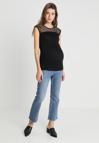 Top de maternidade preto com ombros transparentes em pontos, combinado com jeans claros cropped azuis e mules pretos, apresentando um design ajustado e leve franzido.