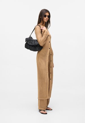 Γυναίκα που φοράει ένα μακρύ μπεζ πλεκτό ζακέτα και παντελόνι, με μαύρα γυαλιά ηλίου, μαύρα σανδάλια και κρατώντας μια μαύρη τσάντα ώμου με βολάν.