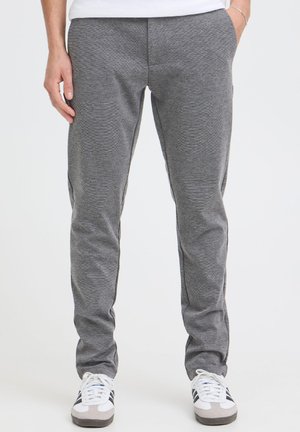Personne portant un pantalon de jogging gris et des baskets blanches à bandes noires, les mains dans les poches, vue de la taille vers le bas sur fond uni.