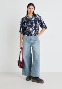 Blusa floreale blu navy con maniche a sbuffo, abbinata a jeans a gamba larga di colore azzurro chiaro. Il modello porta una borsa a tracolla bordeaux. Stivali a caviglia grigi.