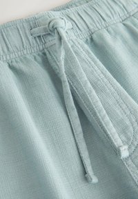 Shorts en tissu bleu clair texturé avec une ceinture élastique et un cordon de serrage noué à l'avant, montrés en détail rapproché.