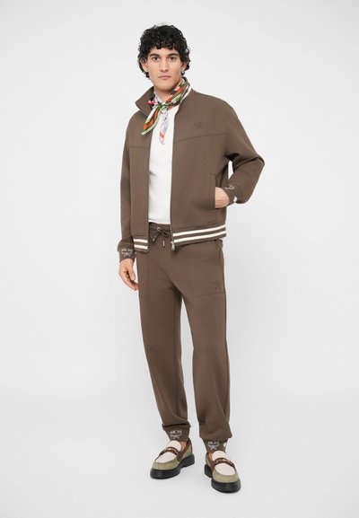 Homem veste um casaco castanho com fecho éclair e calças combinando, camiseta branca, lenço colorido no pescoço e sapatos de dois tons com detalhe em corrente.