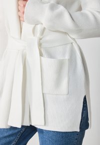 White ribbed cardigan with a waist tie, featuring a front pocket and side slits, paired with blue denim pants.