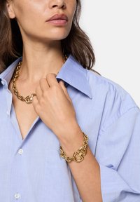 Guldkæde og armbånd med sammenkoblede, overdimensionerede segmenter. Båret med en lyseblå skjorte med krave, der fremhæver de rene linjer og glatte teksturer.