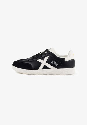 Zapatillas deportivas en blanco y negro con una parte superior de cuero texturizado, suela de goma blanca y un diseño distintivo en forma de cruz en el lateral.
