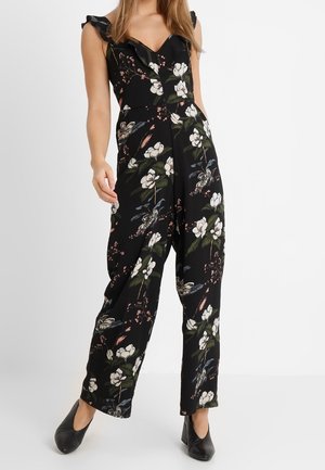 Femme portant une combinaison noire à motifs floraux sans manches avec des bretelles à volants et des chaussures noires à talons.