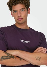 Lilla bomulds T-shirt med rund halsudskæring; har hvid trykt tekst "CURATED STAPLES." Modellen står med armene korset, der viser tatoveringer.