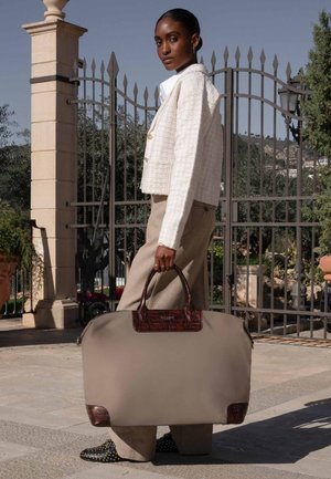 Sac de voyage beige avec des accents en cuir brun texturé, forme arrondie et doubles poignées, porté par une personne en veste légère et pantalons ajustés.
