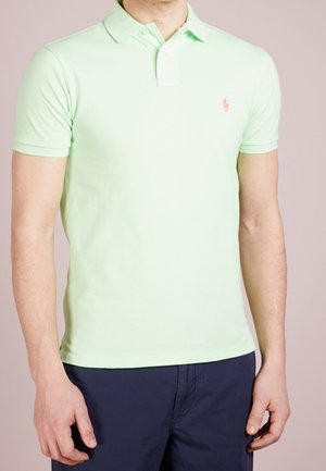 Man in een lichtgroen polo shirt met korte mouwen en kraag, met donkerblauwe broek, staande tegen een effen achtergrond.