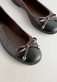 Ballerines en cuir marron avec une coque en pointe noire texturée, ornées d'un nœud en ruban marron et de détails en métal argenté. Doublure intérieure lisse.