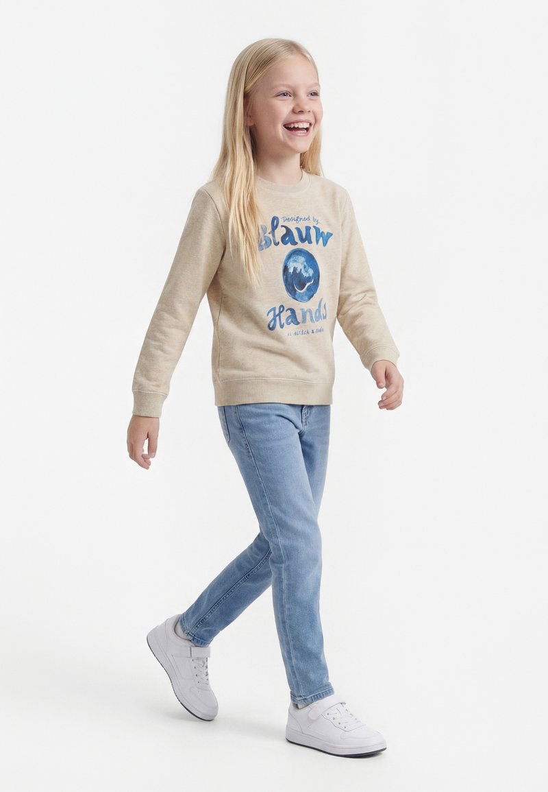 Junges Mädchen geht, lächelt, trägt einen beigen Pullover mit blauem Aufdruck, hellblaue Jeans und weiße Turnschuhe vor weißem Hintergrund.