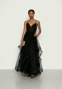 Sieviete, kas ģērbusies melnā bezpiedurkņu tilla kleitā ar pieguļošu augšdaļu, tur rokā bežkrāsas makam, stāvot uz neitrāla fona.