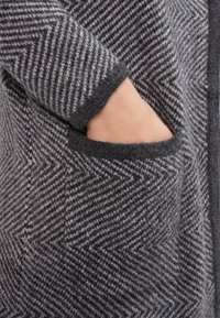 Cárdigan de punto con patrón de espiga en gris y negro, con una textura suave, que incluye un bolsillo lateral y un ribete oscuro contrastante en las mangas.