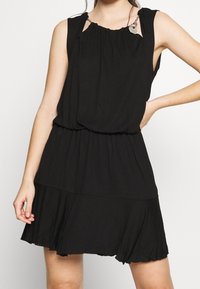 Kobieta w bezrękawowej czarnej sukience z łańcuszkowymi ramiączkami i marszczonym pasem, pozująca z jedną ręką na biodrze na białym tle.