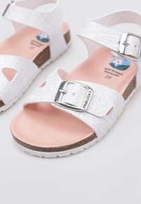 Sandalias blancas texturizadas con correa ajustable, herrajes de hebilla plateada, plantilla contorneada marrón y suela blanca. Ideales para uso informal.