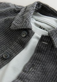Grijze corduroy shirtjack met een gestructureerd oppervlak, voorzien van een knoopsluiting en een lichtgrijze binnenlaag. Zwarte knopen met een glanzende afwerking.