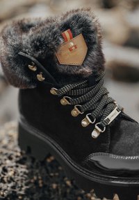 Botín negro de ante con un cuello de pelaje sintético, herrajes dorados, cordones texturizados y una puntera brillante. Presenta un acento de cuero con marca.