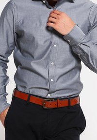 Šedá košile s hladkým povrchem, nošená nepřičepovaná přes tmavé kalhoty, doplněná hnědým koženým páskem s kovovou sponou.