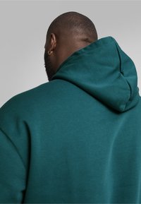 Sudadera color verde azulado con capucha ajustable, hecha de un tejido suave. Presenta un corte holgado, costuras visibles y una textura suave.