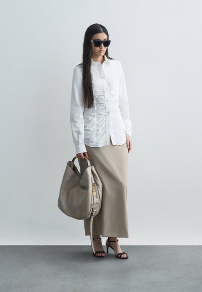 Sieva, tērpta baltā drapētā blūzē, bēšā garajā svārku, melnās augstpapēžu sānēs, melnās saules brillēs, rokās tur lielu bēšas krāsas rokassomu ar zelta logotipa detaļām.