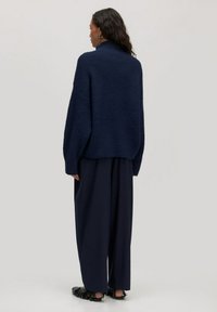 Navy gestrickter Oversized-Pullover mit hohem Kragen, sanfter Textur und überschnittenen Schultern, kombiniert mit weiten, dunklen Hosen und flachen Schuhen.