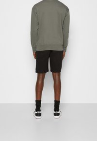 Persona che indossa una felpa verde oliva, pantaloni corti neri, calze nere e sneaker bianche Vans, in piedi contro uno sfondo bianco semplice.