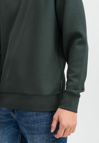 Mørkegrønn hettegenser med glatt tekstur, med rund hals og ribbestrikkede ermer, båret over blå denimjeans.