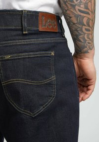 Jeans in denim blu scuro presentano una patta in pelle marrone con la scritta "Lee" in rilievo, cuciture gialle e un design della tasca posteriore.