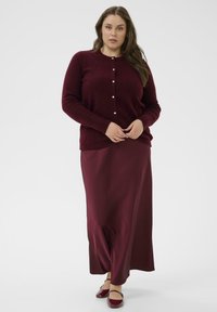 Bordo cardigan z gumbom, v kombinaciji z dolgo satensko bordo krilo in zaprto-točilnimi čevlji; mehka tekstura in prava oblika.