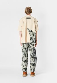 Persona con cabello corto y ondulado que lleva una camisa oversized beige con un parche estampado y pantalones texturizados a juego con largas etiquetas en blanco y negro, zapatos marrones.