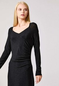 Femme blonde portant une robe noire cintrée à manches longues avec un décolleté carré, se tenant devant un fond gris clair uni.