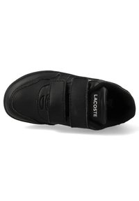 Fekete sportcipő tépőzáras pántokkal, sima szintetikus felsőrésszel, gumitalppal, és az oldalon található "LACOSTE" logóval. Kerek orrú kialakítás.